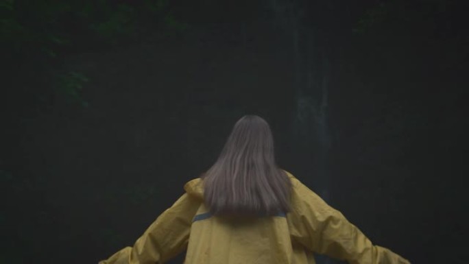 年轻女孩旅行者徒步旅行者穿着黄色雨衣奔向瀑布，举起双手，享受自然和生活