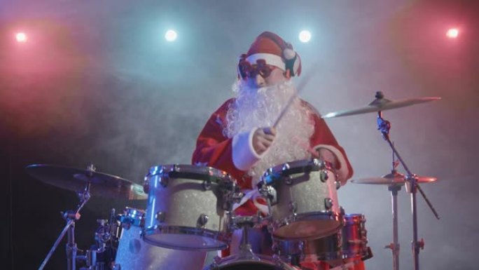 穿着红色服装的圣诞老人留着胡须，戴着滑稽的眼镜，在黑暗的工作室里打鼓，灯光明亮，烟雾缭绕。一个穿着狂
