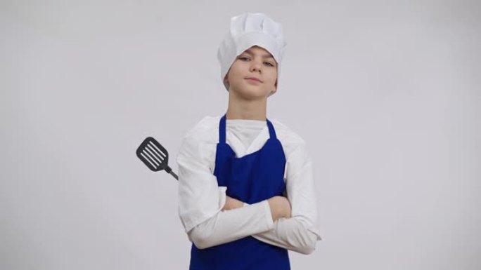 自信的小厨师与开槽的特纳在白色背景下摆姿势。戴着炊事帽和围裙的高加索男孩的肖像用严肃的面部表情看着相