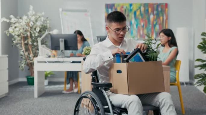 年轻的公司办公室工作人员戴着眼镜受伤，事故，残疾，坐在轮椅上，从桌子上举起一盒用具，退休，下岗，失业