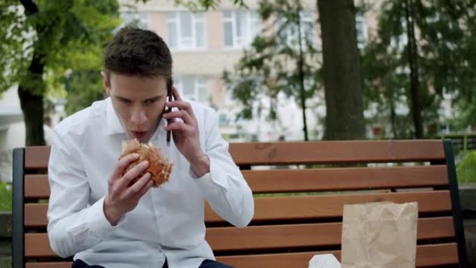 穿着西装的饥饿男子在板凳上微笑着在电话上聊天时吃汉堡