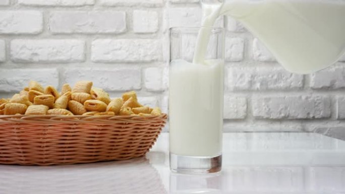 将新鲜的乳制品倒入白色砖墙背景下的玻璃杯中，新鲜的牛奶和糕点放在厨房的桌子上。从水罐里倒牛奶。奶昔