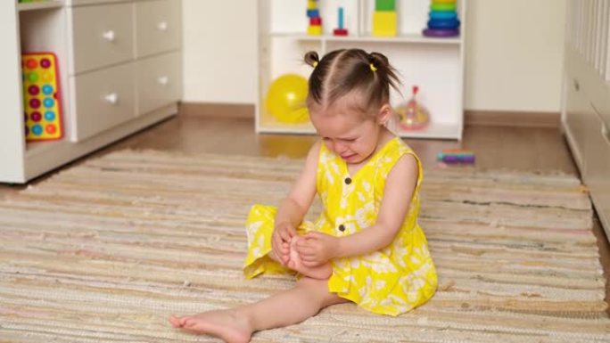 一个穿着裙子的两岁女孩正在哭泣，手指着脚趾，坐在托儿所的地板上。