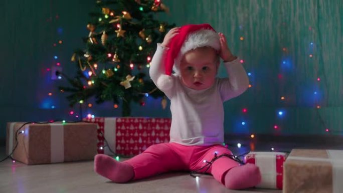 一个有趣的小女孩坐在圣诞树旁，脱下圣诞帽。慢动作。寒假的概念