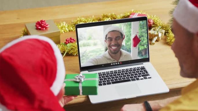 高加索人的父亲和儿子戴着圣诞老人的帽子，使用笔记本电脑进行圣诞节视频通话，屏幕上有男子