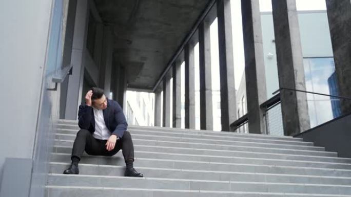 年轻的亚洲沮丧男子坐在办公室商务中心大楼附近的楼梯户外街道上