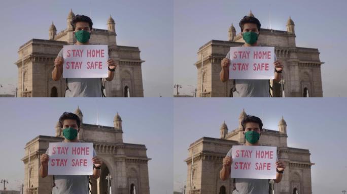 一个年轻自信的男人或戴着口罩的男性站立并举着标语牌，上面写着 “待在家里保持安全”。