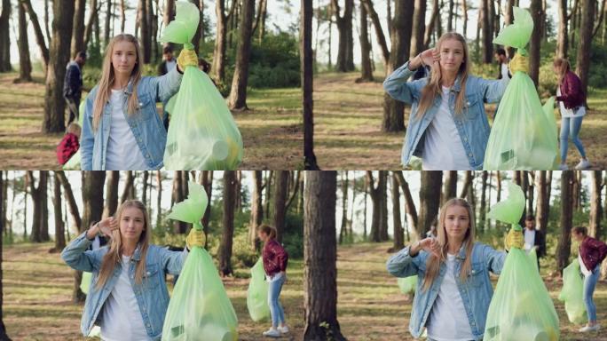 漂亮活泼的年轻金发女郎拿着垃圾袋，在镜头上显示出她不喜欢和其他市民一起捡垃圾——公园的志愿者，正面
