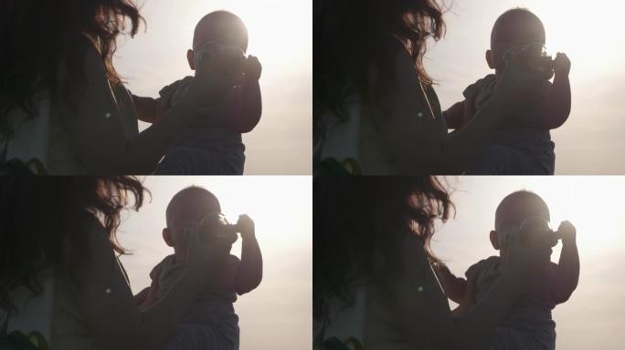 母亲在户外散步时从婴儿奶瓶中给小婴儿水，可爱的高加索人六个月大的男婴在蓝天和阳光的背景下背光中喝淡水