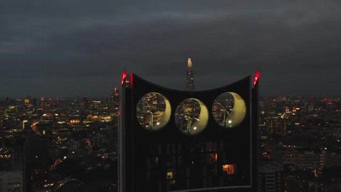 在分层公寓楼的照明顶部飞来飞去。三台风力涡轮机集成到房屋建筑中。夜市景观的鸟瞰图。英国伦敦