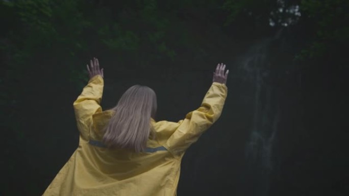 年轻女孩旅行者徒步旅行者穿着黄色雨衣奔向瀑布，举起双手，享受自然和生活