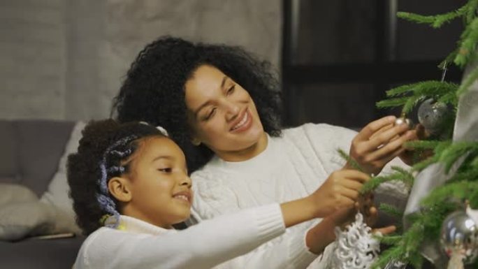 快乐的非洲裔美国妈妈和小女儿的肖像用节日玩具装饰圣诞树，同时坐在阁楼风格的家庭房间的背景下。特写。慢