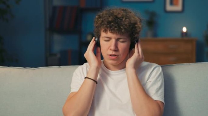 一个卷发的少年坐在客厅的沙发上，耳朵上戴着无线耳机，双手放在上面。这位少年正在听音乐，接受歌曲的歌词