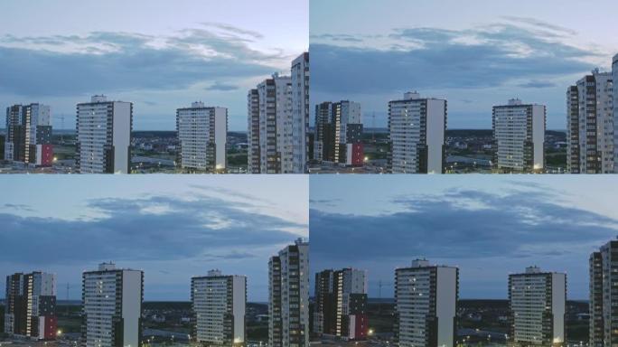 黄昏城市上空的云的延时视频，城市中有公寓的多层建筑和一层住宅，城市上空的夜幕降临，窗户上的灯点亮