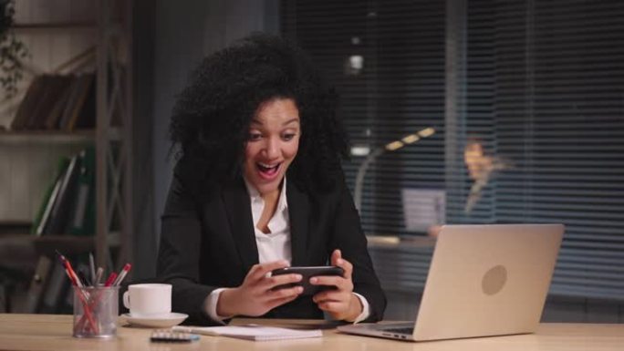 非裔美国妇女在智能手机上发短信的肖像，很高兴收到好消息，庆祝胜利。女商人在工作场所摆姿势，坐在办公室