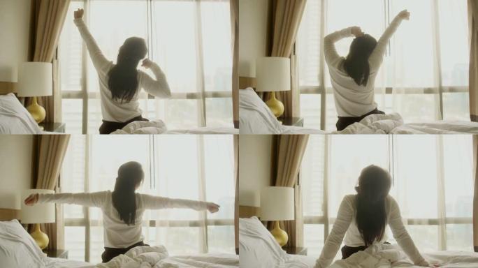 年轻清新的亚洲女女人早上醒来穿着睡衣在床上升起手，在阳光照射下的大窗户旁边，covid锁定结束后的假