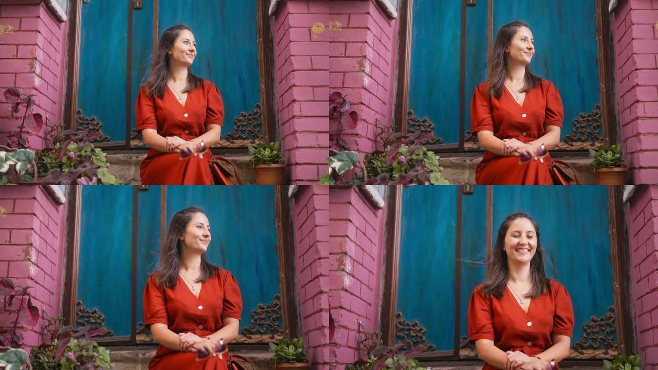 穿着红色长裙的美女坐在台阶上，在城市里对着镜头微笑