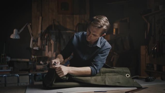 工匠爱好。年轻专注的人裁缝在他的车库车间里挑选旧帆布包，用刀切割接缝