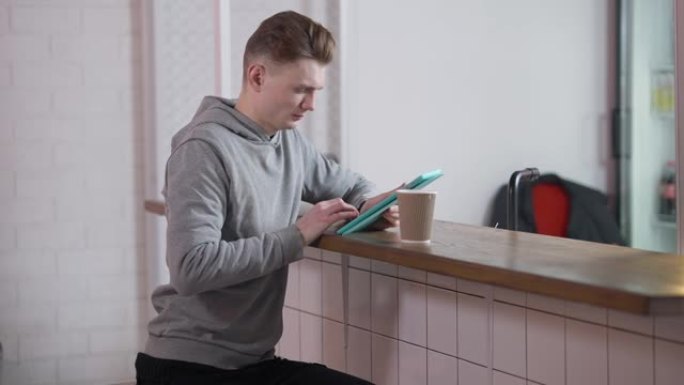 吸收千禧一代高加索年轻人坐在柜台在咖啡馆的柜台在平板电脑应用程序在线消息。集中自信的学生在食堂上网。