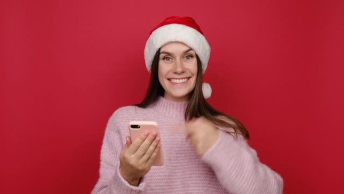 穿着粉色毛衣和圣诞帽的可爱漂亮年轻圣诞白人妇女用手机打字说哇是的显示拇指向上像手势，在红色背景工作室