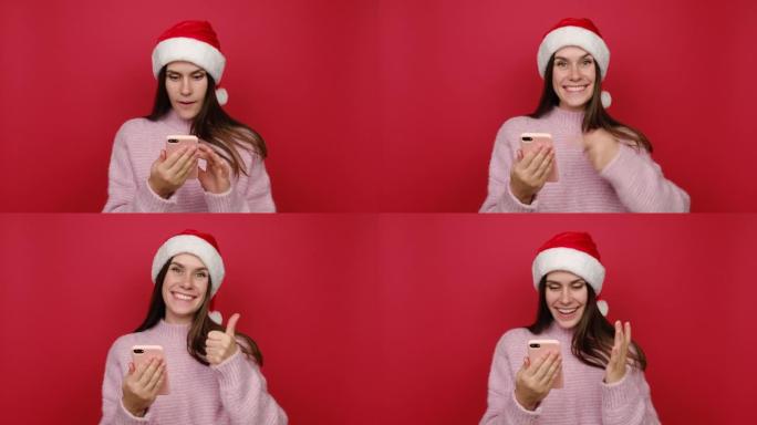 穿着粉色毛衣和圣诞帽的可爱漂亮年轻圣诞白人妇女用手机打字说哇是的显示拇指向上像手势，在红色背景工作室