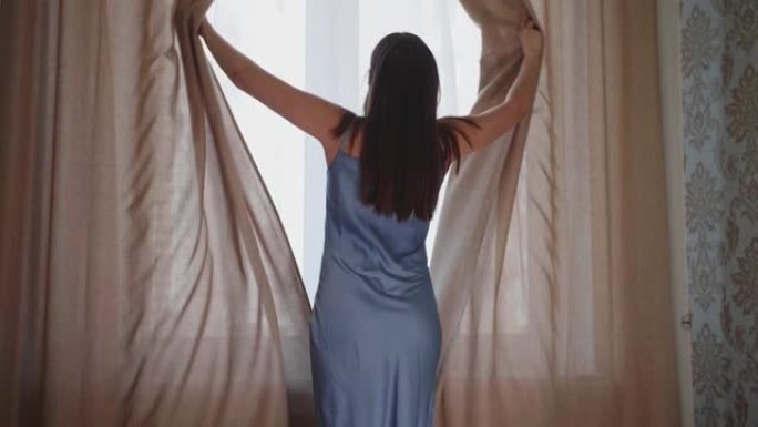年轻女子走近卧室的窗户，用手推米色窗帘，享受黎明。她留着深色长发，穿着蓝色衬衫。早晨，休息，新的一天