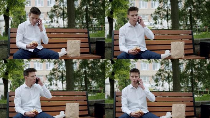 穿着西装的男人坐在智能手机上聊天，坐在街头长凳上吃汉堡