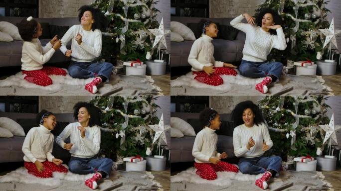 非裔美国妈妈和她的小女儿滑稽跳舞和微笑的肖像。女人和女孩坐在沙发和装饰圣诞树附近。家庭之夜快乐。慢动