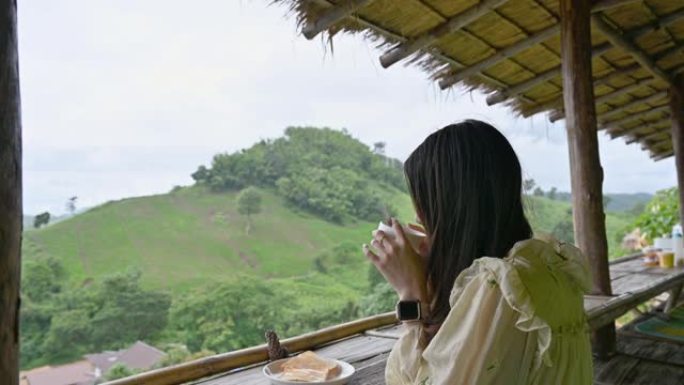 年轻的亚洲妇女早上在山间的小屋阳台上喝热巧克力和烤面包