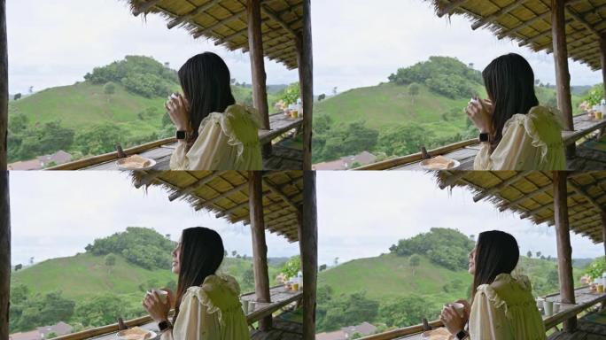 年轻的亚洲妇女早上在山间的小屋阳台上喝热巧克力和烤面包