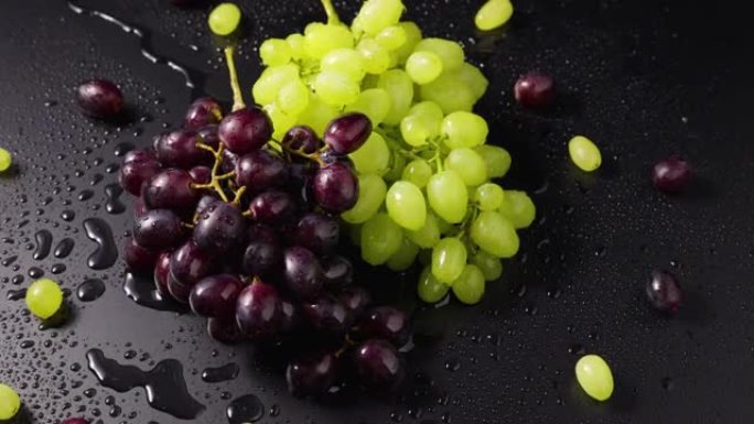 一束绿色和蓝色的葡萄，湿漉漉的水滴躺在潮湿的桌子表面上。慢动作旋转的成熟葡萄浆果的俯视图特写。水滴葡