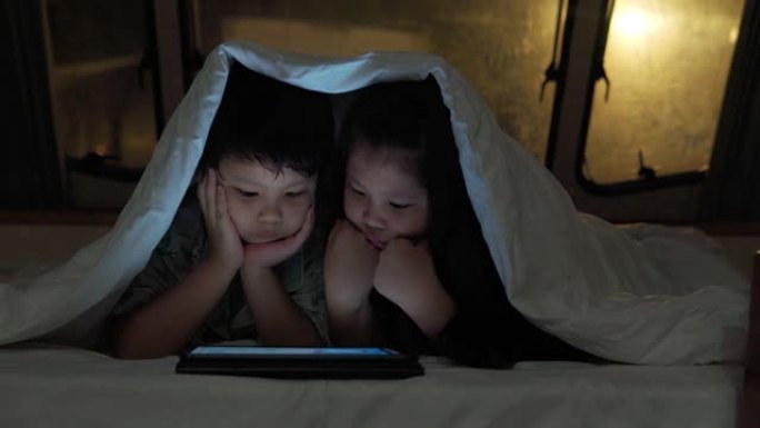 亚洲兄弟姐妹在大篷车的毯子下使用平板电脑。当他们晚上躺在前面时。房车假日旅行