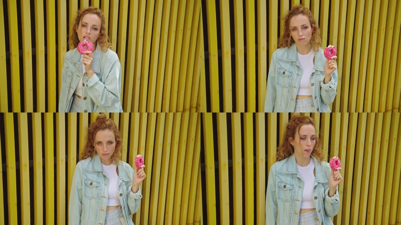 红色卷发年轻女子吃粉红色甜甜圈的肖像。穿着牛仔裤夹克的性感女孩靠在黄色的木栅栏上。漂亮的姜咬甜甜圈食