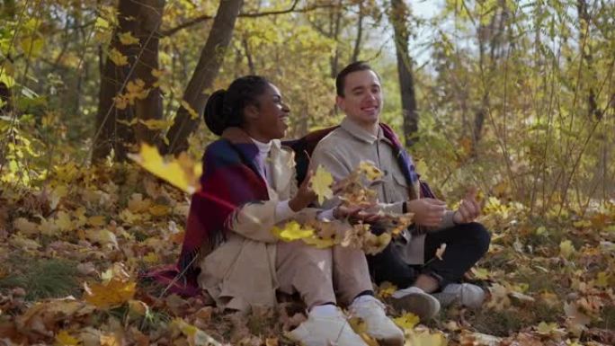 混合赛跑的夫妇在秋天坐在公园的毯子上，向空中扔树叶