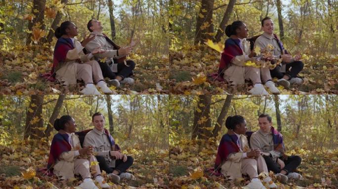 混合赛跑的夫妇在秋天坐在公园的毯子上，向空中扔树叶