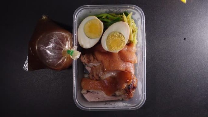 猪腿饭装在一个塑料盒中，上面放着蘸酱。黑桌上的肉汤和大蒜辣椒粉，泰国菜