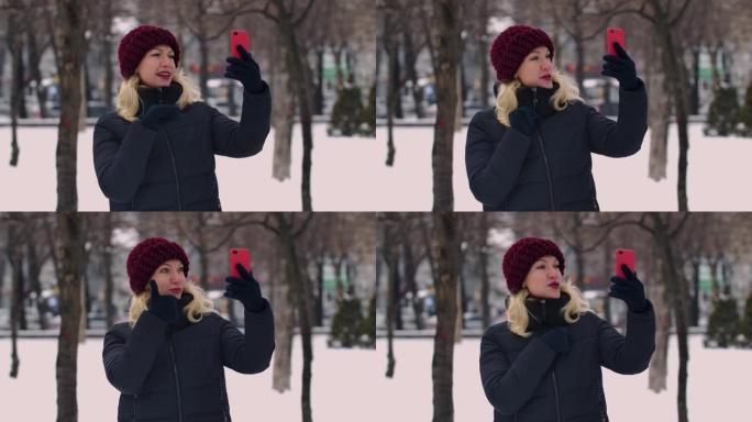 一个冬天白雪皑皑的公园里的女人拿着智能手机，通过视频通话。女孩向对话者打招呼，显示拇指向上的手势，发