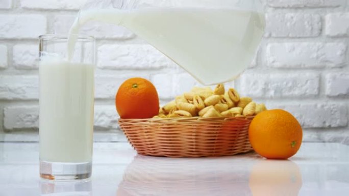将水罐中的牛奶倒入玻璃杯中，在白色的厨房桌子上放着水果和糕点的牛奶静物。在白色砖墙背景上倒牛奶