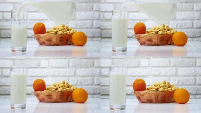 将水罐中的牛奶倒入玻璃杯中，在白色的厨房桌子上放着水果和糕点的牛奶静物。在白色砖墙背景上倒牛奶