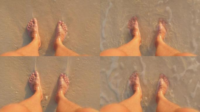 站在飞溅的冲浪中的沙滩上的男性脚的第一人称视角。正上方可见