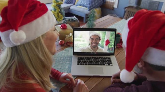 微笑的高加索夫妇戴着圣诞老人的帽子，使用笔记本电脑与屏幕上的男人进行圣诞节视频通话
