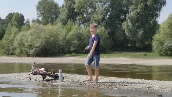 可爱的高加索少年赤脚站在河岸上向水中扔石头。大自然中的休闲活动。慢动作。