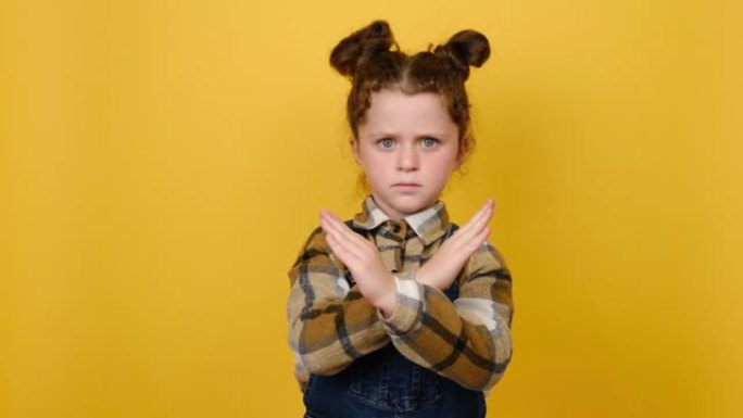 肖像不高兴的小女孩，手握手掌折叠双手，以停止手势，穿着衬衫，在工作室的黄色背景墙上孤立地摆姿势。童年