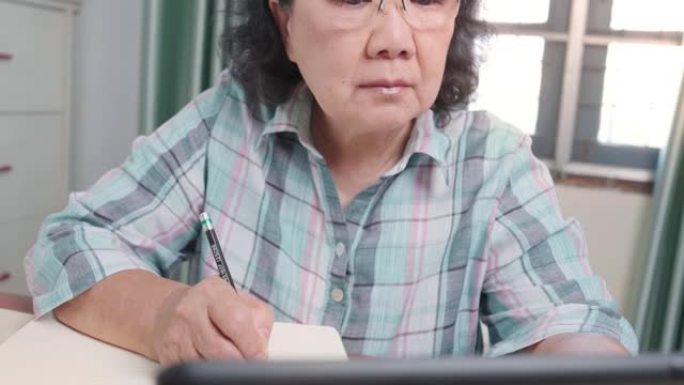 亚洲高级女性戴眼镜在无线平板电脑上观看在线课程，在线学习，老年人在空闲时间，在笔记本上写讲座，在家，