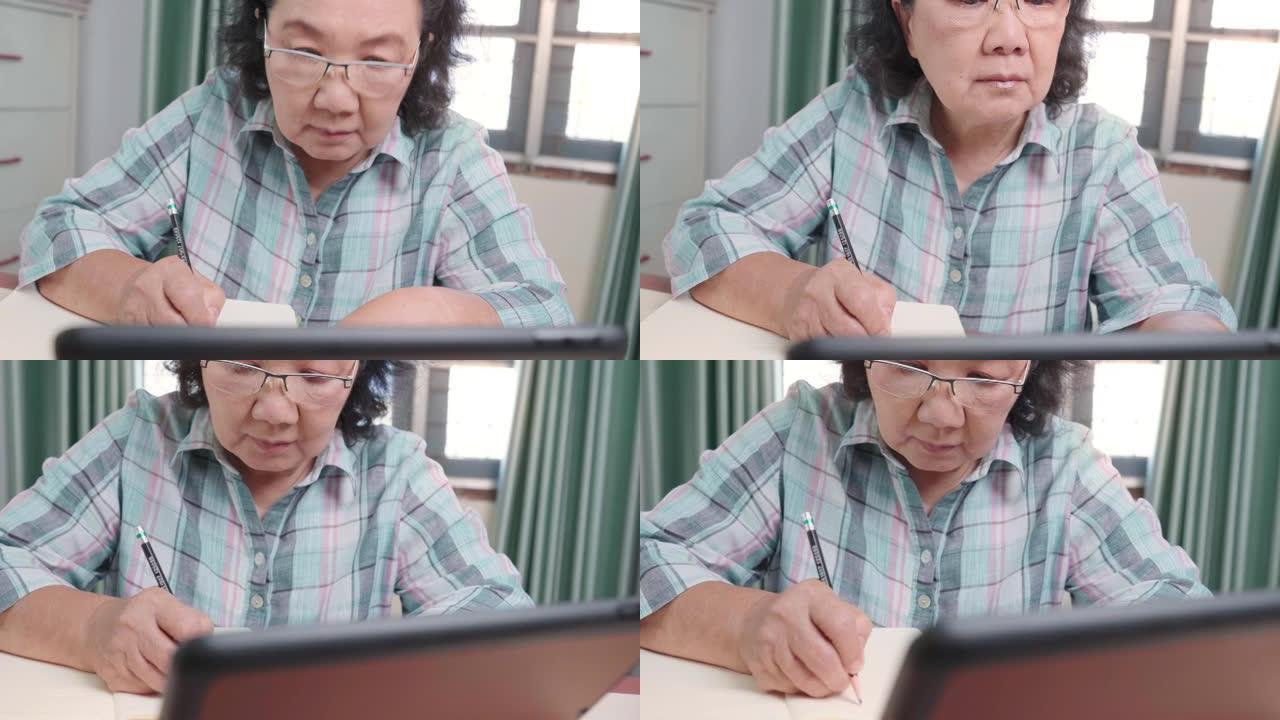亚洲高级女性戴眼镜在无线平板电脑上观看在线课程，在线学习，老年人在空闲时间，在笔记本上写讲座，在家，
