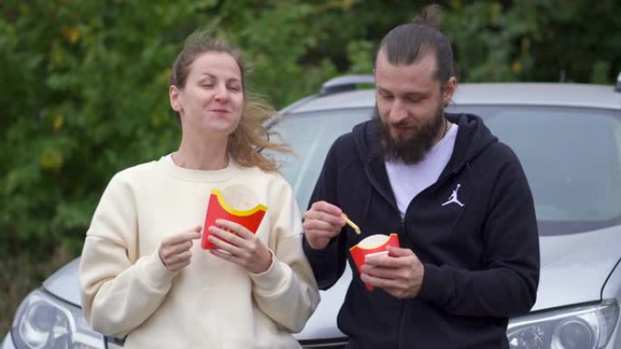 一对夫妇在公园约会时一起吃麦当劳薯条。垃圾食品浪漫小吃。森林里的绿树。青少年在车上吃零食。说笑。快乐