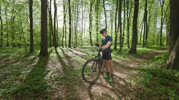 一名骑自行车的人站在森林中间，看着智能手机上的训练数据。他旁边有一辆自行车。他穿着自行车制服。4K 