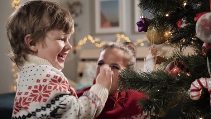 孩子正在装饰圣诞树。快乐的孩子把玩具放在圣诞树的树枝上，对这一刻感到非常高兴，背景中的母亲看着婴儿并