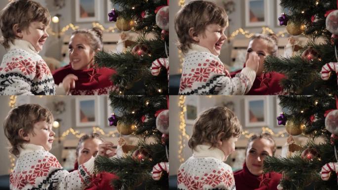 孩子正在装饰圣诞树。快乐的孩子把玩具放在圣诞树的树枝上，对这一刻感到非常高兴，背景中的母亲看着婴儿并
