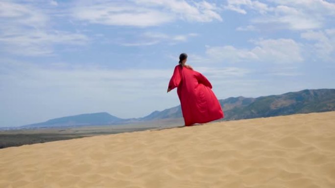 穿着红色缎子长裙的性感黑发女人走在沙漠中的沙丘上